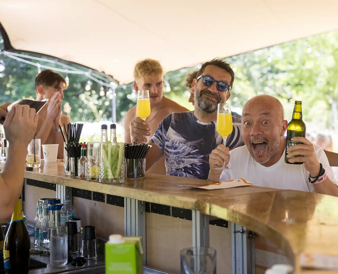 Two men at a bar raising drinks, one holding a beer bottle and the other a glass of orange cocktail.