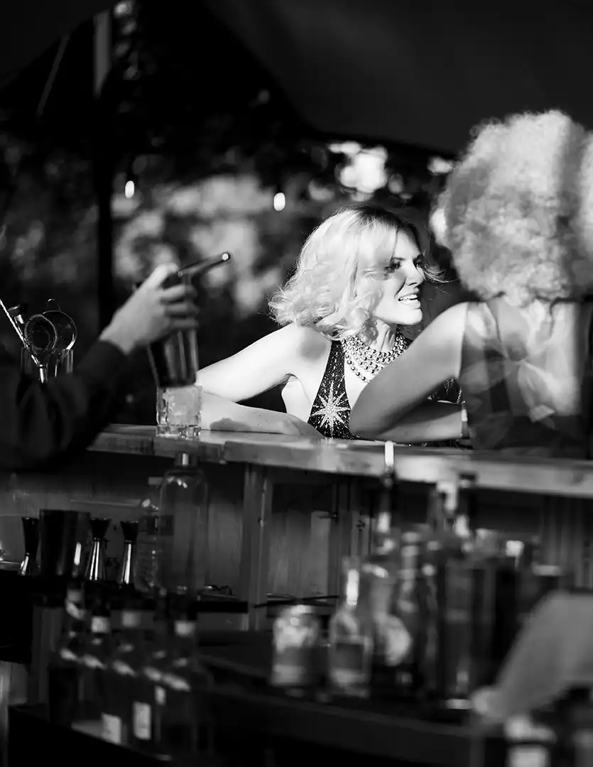 Two women at a bar talking, one smiling, with a bartender holding a shaker nearby.