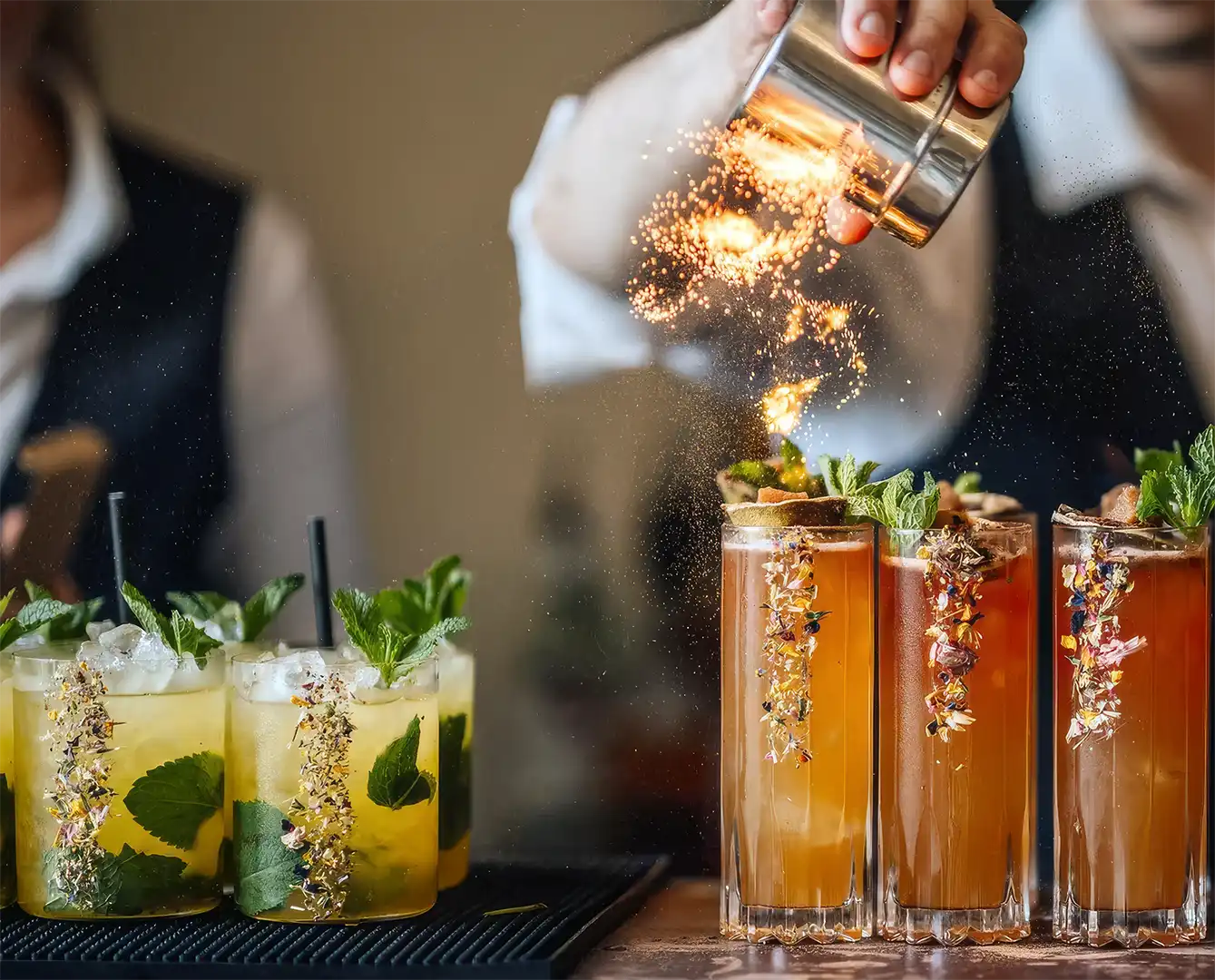 Bartender sprinkles garnish onto six decorated cocktails, three yellow and three amber with mint leaves.