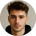 Portrait of a young man with short brown hair and light stubble wearing a black hoodie against a neutral background.