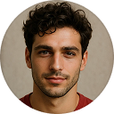 Portrait of a young man with curly dark hair and a slight smile wearing a red shirt against a neutral background.