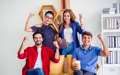 Four young adults enthusiastically raising their fists in a bright room with bookshelves and hexagonal wall shelves.