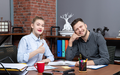 Two young professionals sitting at a desk with documents, smiling, one giving a thumbs-up and the other mimicking a phone gesture.