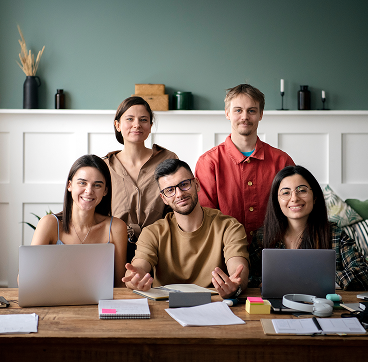 Group of five diverse young adults seated and standing behind a table with laptops, notebooks, and office supplies in a modern workspace.