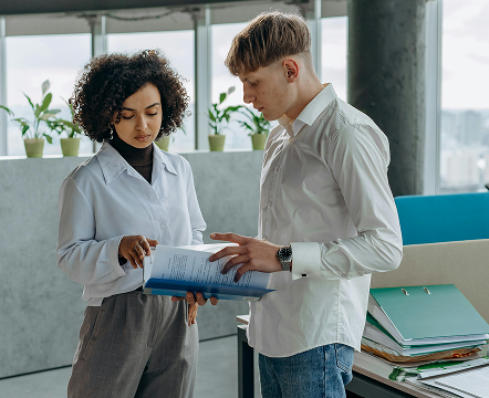 Two coworkers standing and discussing a document in a modern office space with plants and binders in the background.