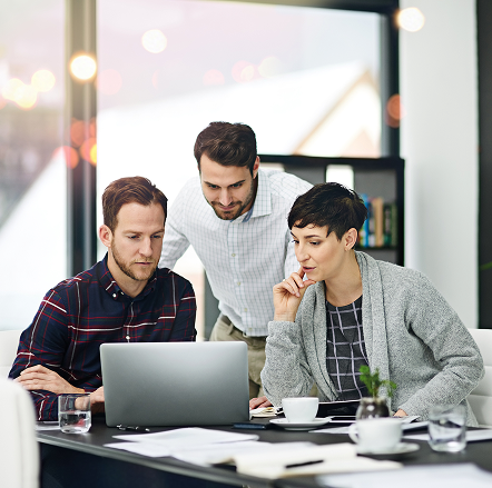 Three coworkers collaborating and looking at a laptop screen in a bright office meeting room.