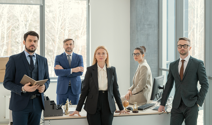 Five professionally dressed businesspeople posing confidently in a modern office with large windows.