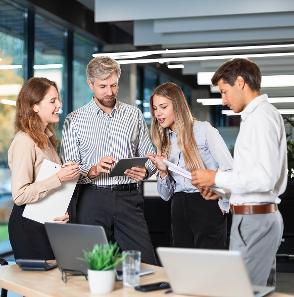 Four colleagues standing and discussing work together in a modern office with laptops and documents on the table.