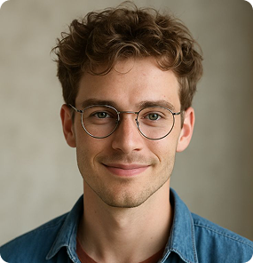Young man with curly brown hair, wearing round glasses and a blue denim shirt, smiling gently at the camera.