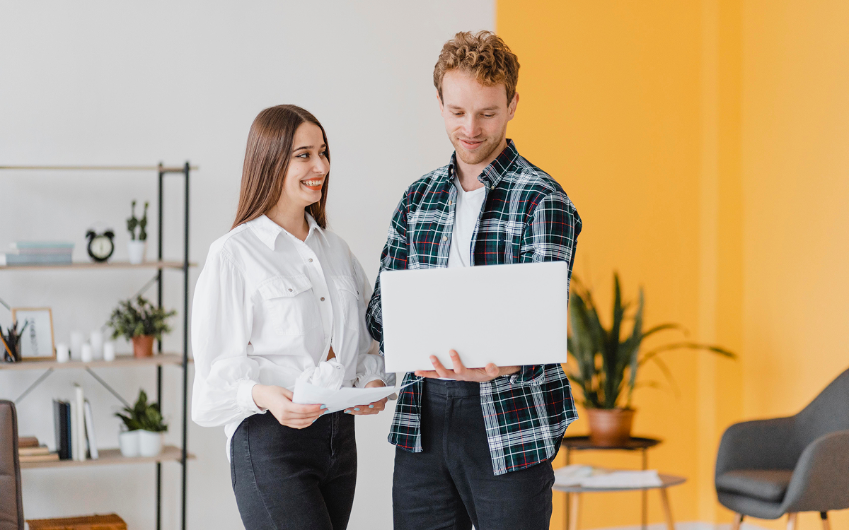 Man holding a laptop and showing something on the screen to a smiling woman holding papers in a bright office.