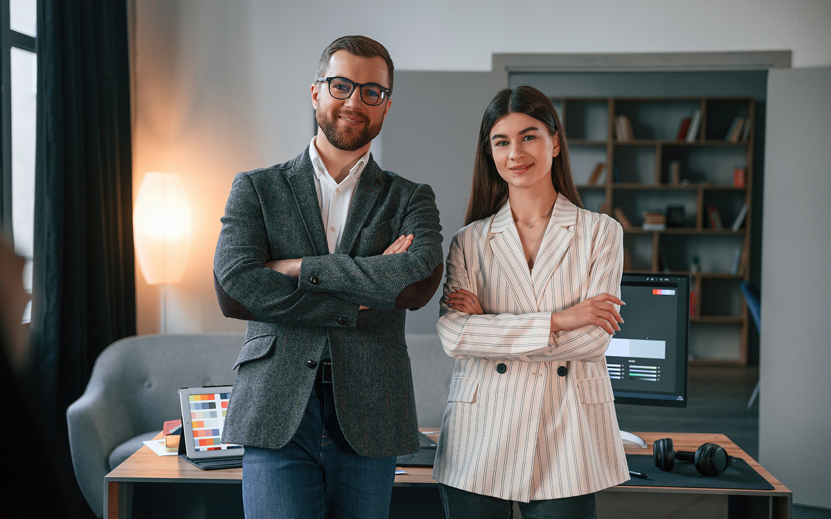 Man and woman standing with arms crossed in an office with computer monitors and a bookshelf behind them.
