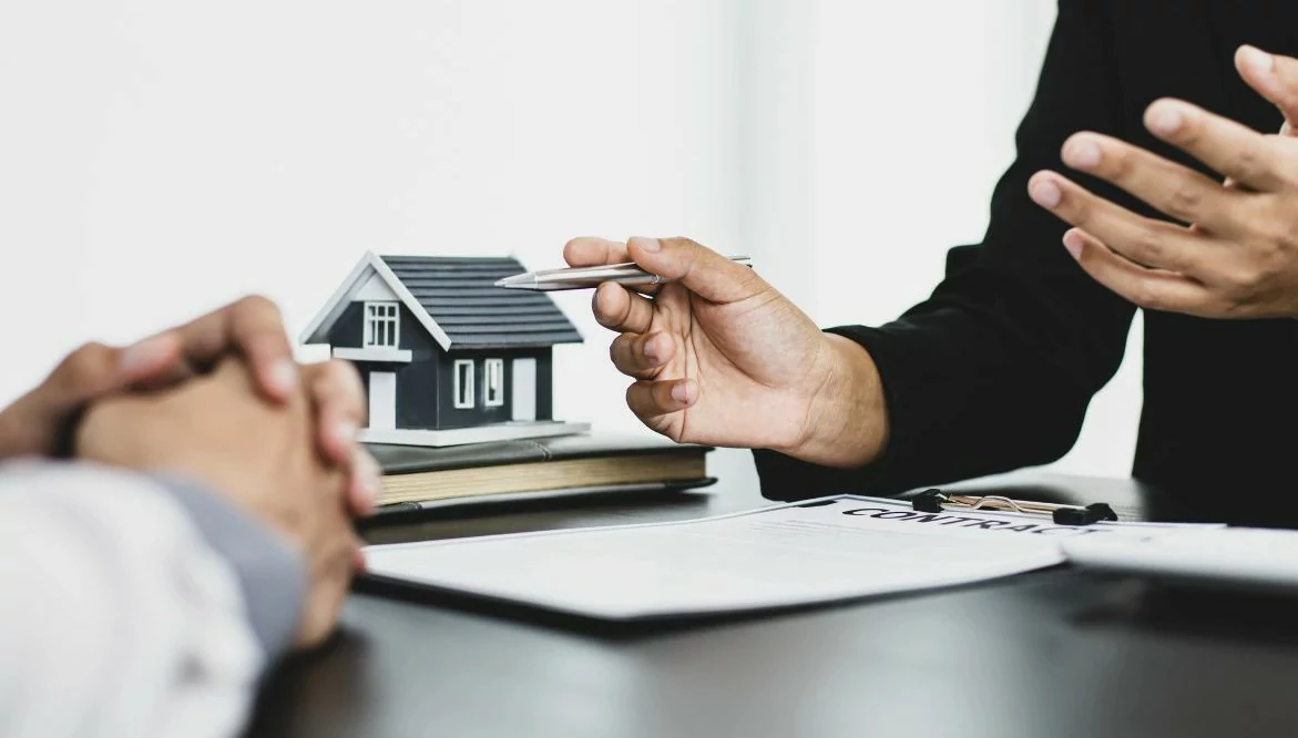 Professionnel immobilier tenant un stylo devant un homme, des documents et une marquette de maison