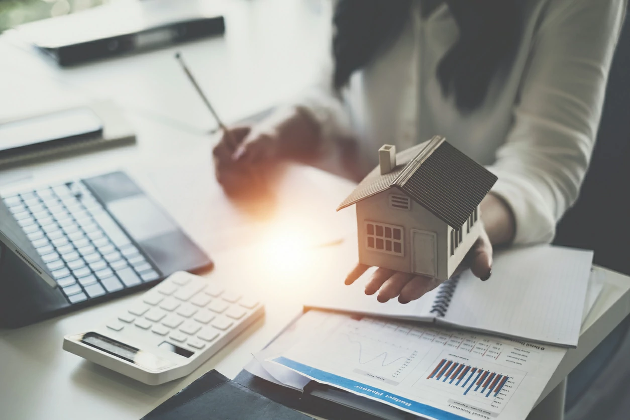 Femme tenant une maquette de maison dans une main et un crayon dans l'autre devant des documents, une calculatrice et un ordinateur portable.