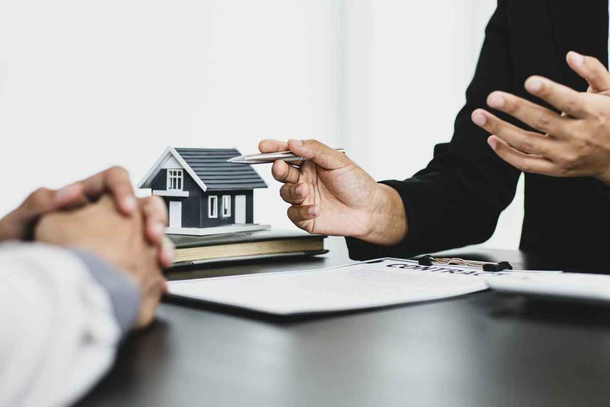 Un professionnel de l'immobilier devant des documents, une maquette de maison et un homme avec les mains croisées.