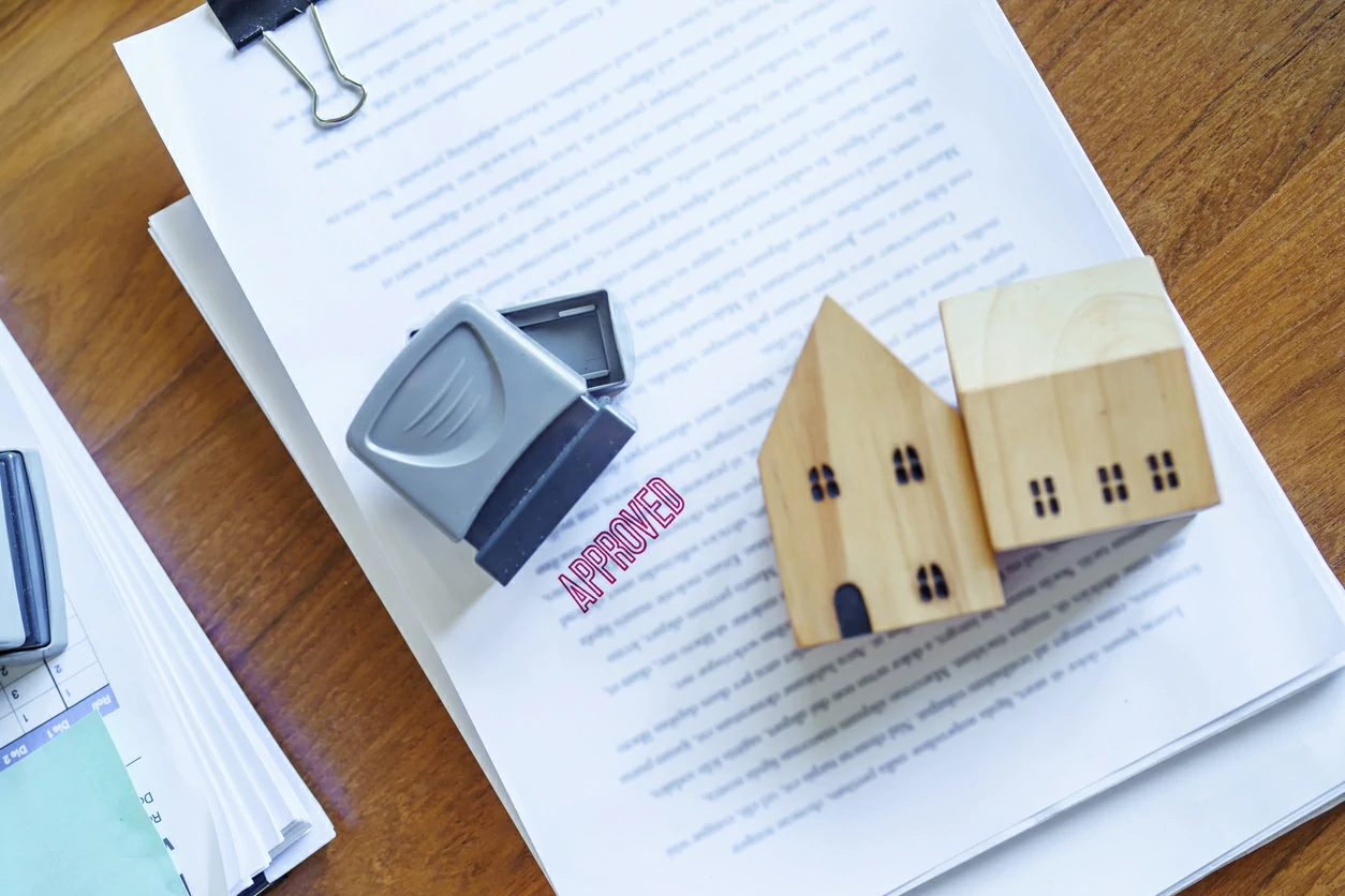 Deux petites maisons en bois et un cachet posés sur une pile de documents.