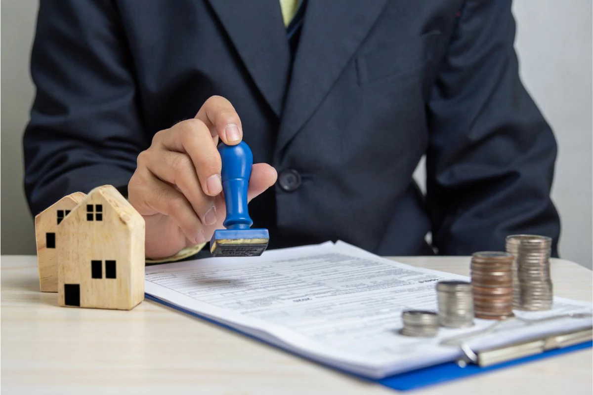 Homme qui tamponne un document avec un cachet à côté de petite maison en bois et des pièces de monnaie.