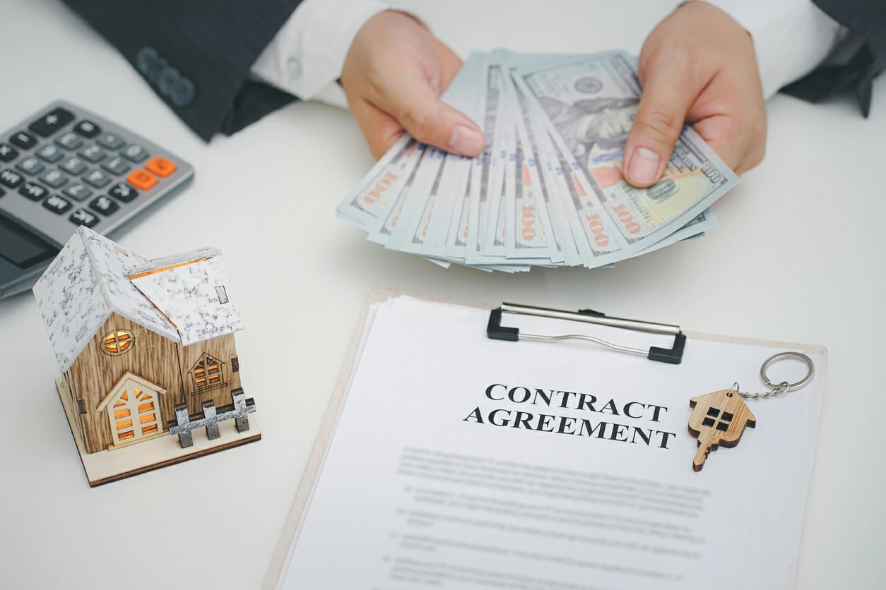 Contrat, calculatrice, petite maison et main tenant des liasses de billets en dollars.