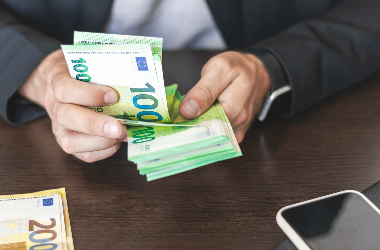 Homme comptant une liasse de billets de 100 euros à la main à côté d'un smartphone.