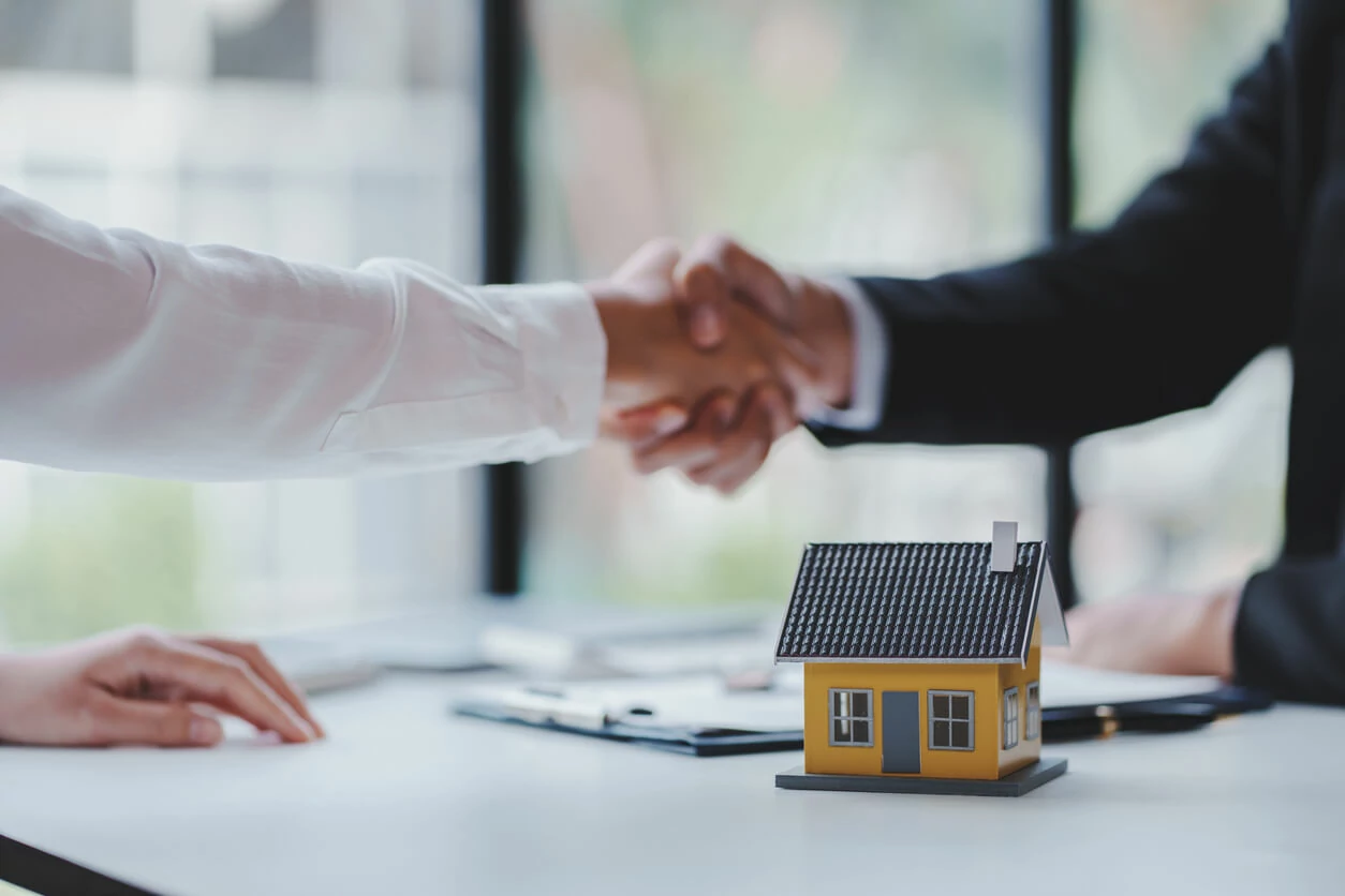 Deux homme se serrant la main au-dessus d'une maison décorative et d'un document.