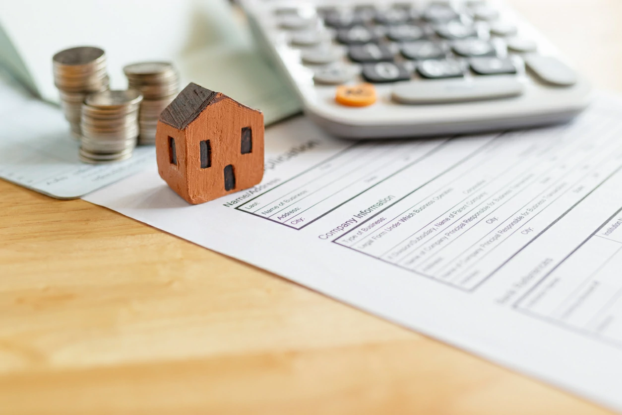 maison miniature en bois, des pièces de monnaie et une calculatrice, le tout posé sur un document.