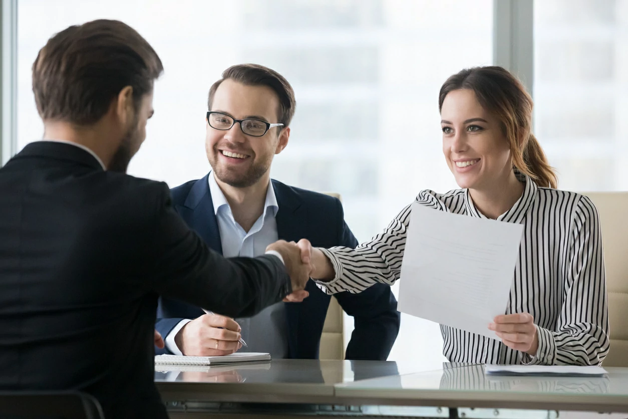 Femme tenant un document et serrant la main d'un professionnel devant elle.