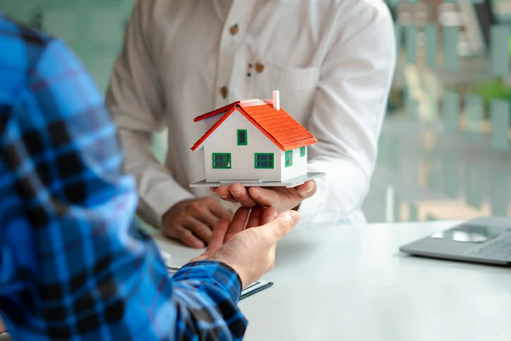 Homme tenant en main une maquette de maison devant un autre homme avec les mains tendues.
