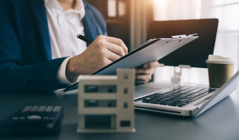 Homme écrivant sur un document à côté d'une maquette d'immeuble et un ordinateur portable.