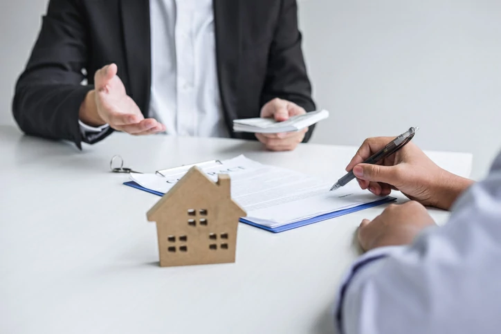 Main signant un document devant une personne tenant une liasse de billet à côté d'une maison en bois.