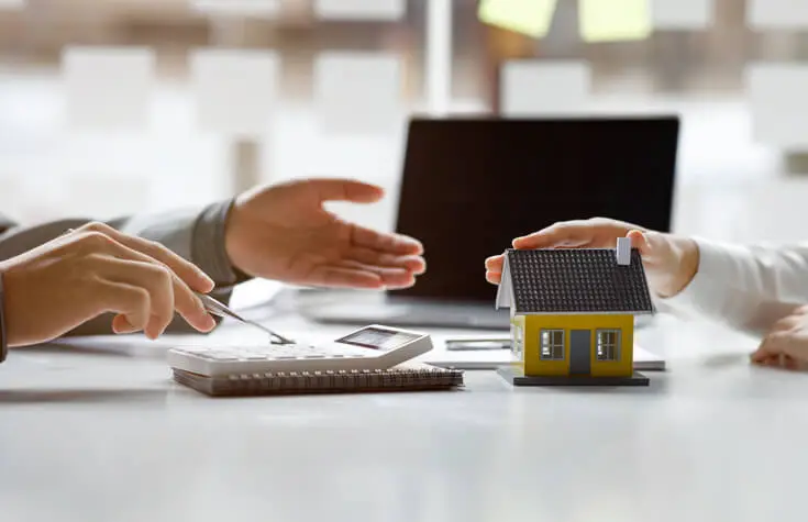 Homme effectuant des calculs avec une calculatrice devant une autre personne avec une main posée sur une petite maison.