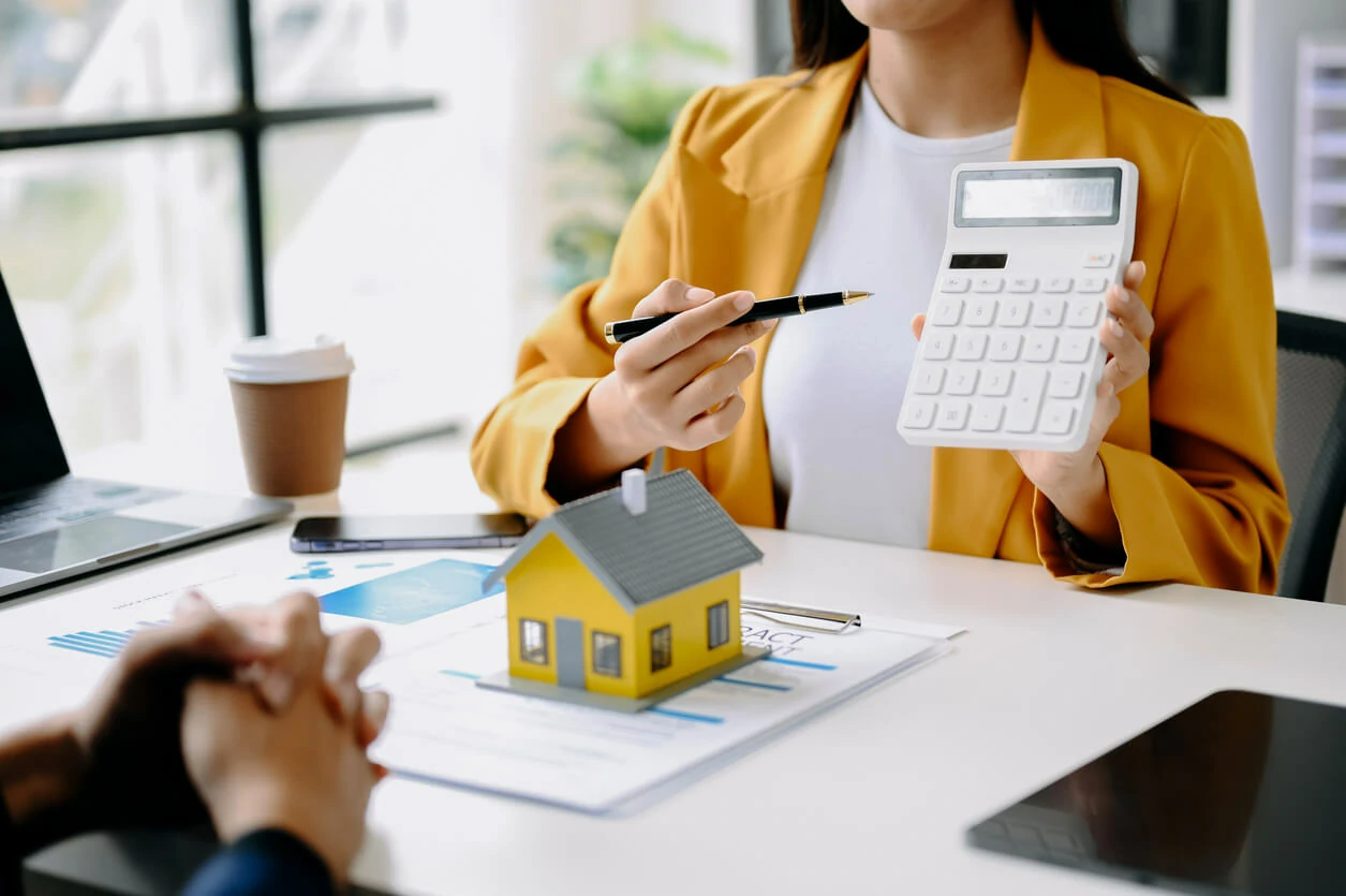 Femme pointant une calculatrice avec un stylo en face d'un homme, de documents et une petite maison.