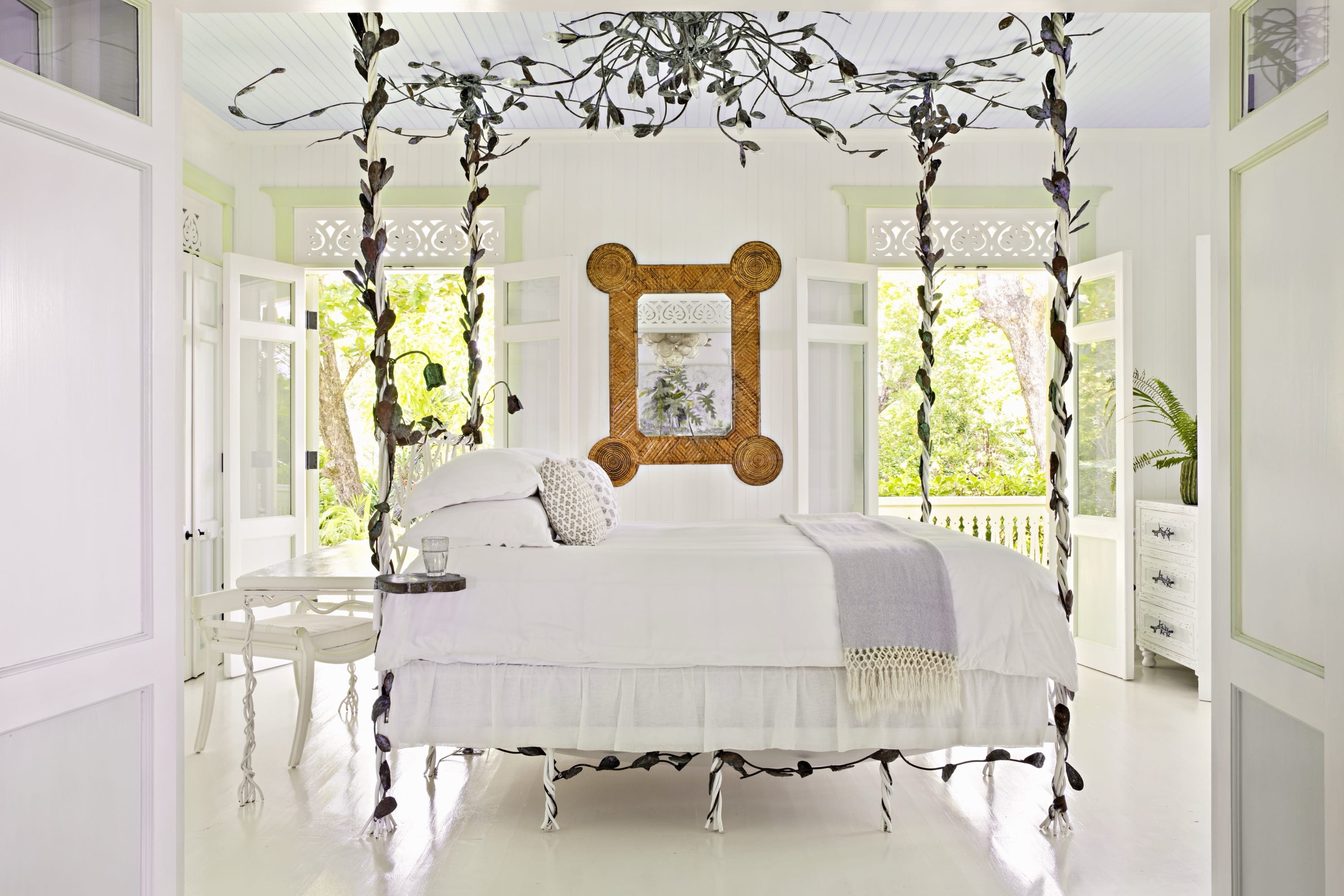 Bright white bedroom with ornate four-poster bed, open windows, wooden mirror, and green plants outside.