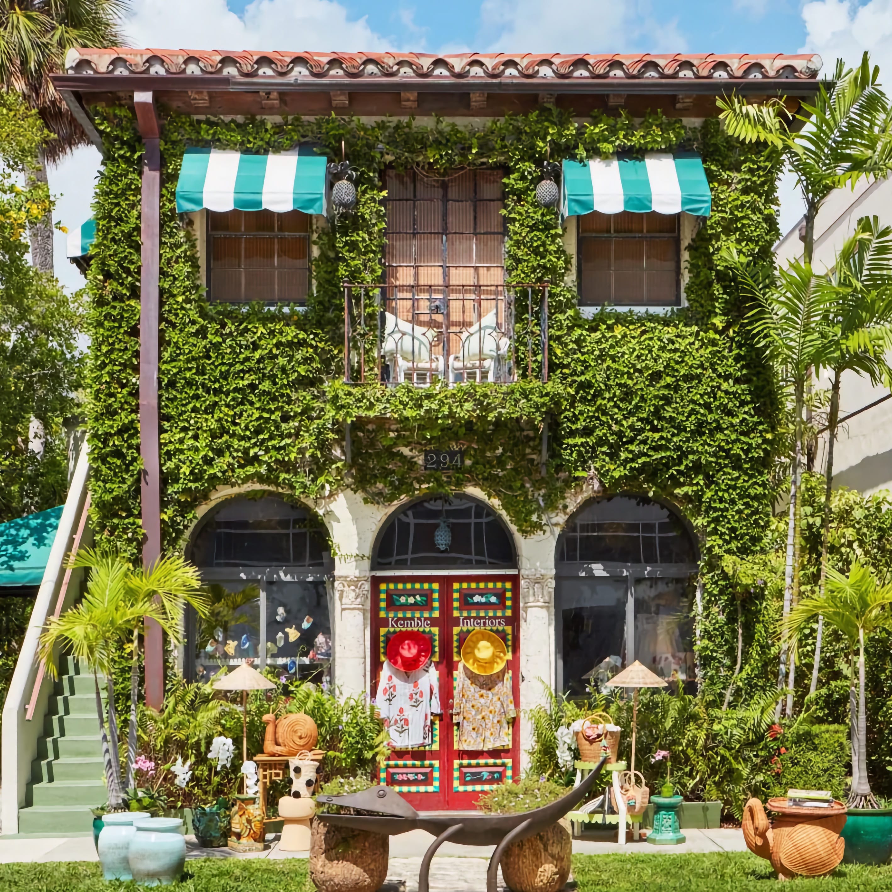 Green ivy-covered storefront with striped awnings, red door, and tropical plants outside.