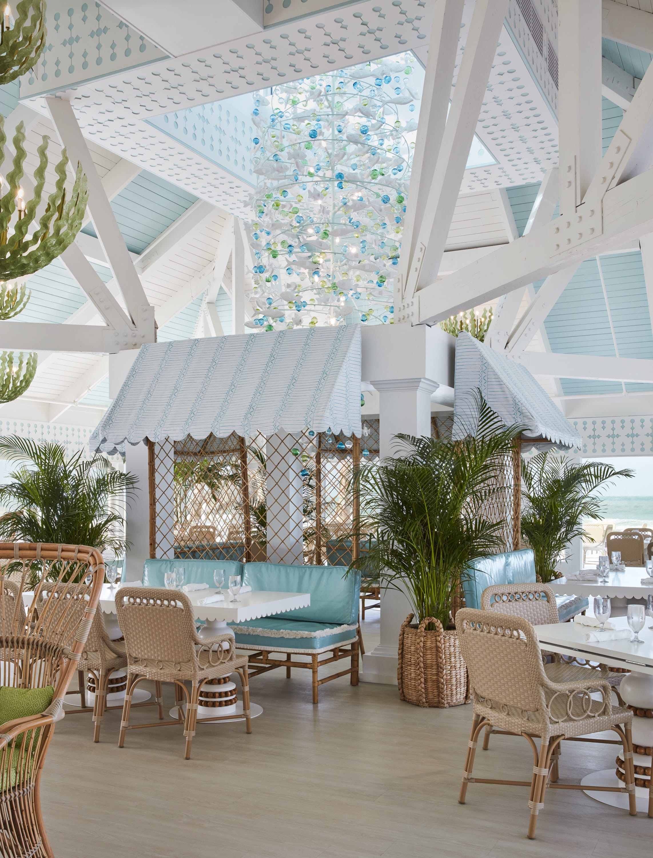 Bright coastal-style restaurant with wicker chairs, turquoise cushions, palm plants, and decorative ceiling lights.