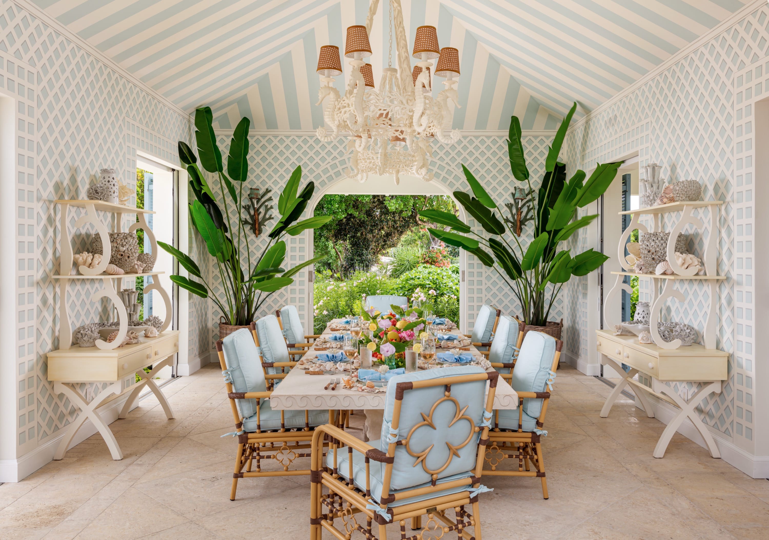 Elegant dining room with blue cushioned chairs, large plants, patterned walls, and a floral centerpiece.