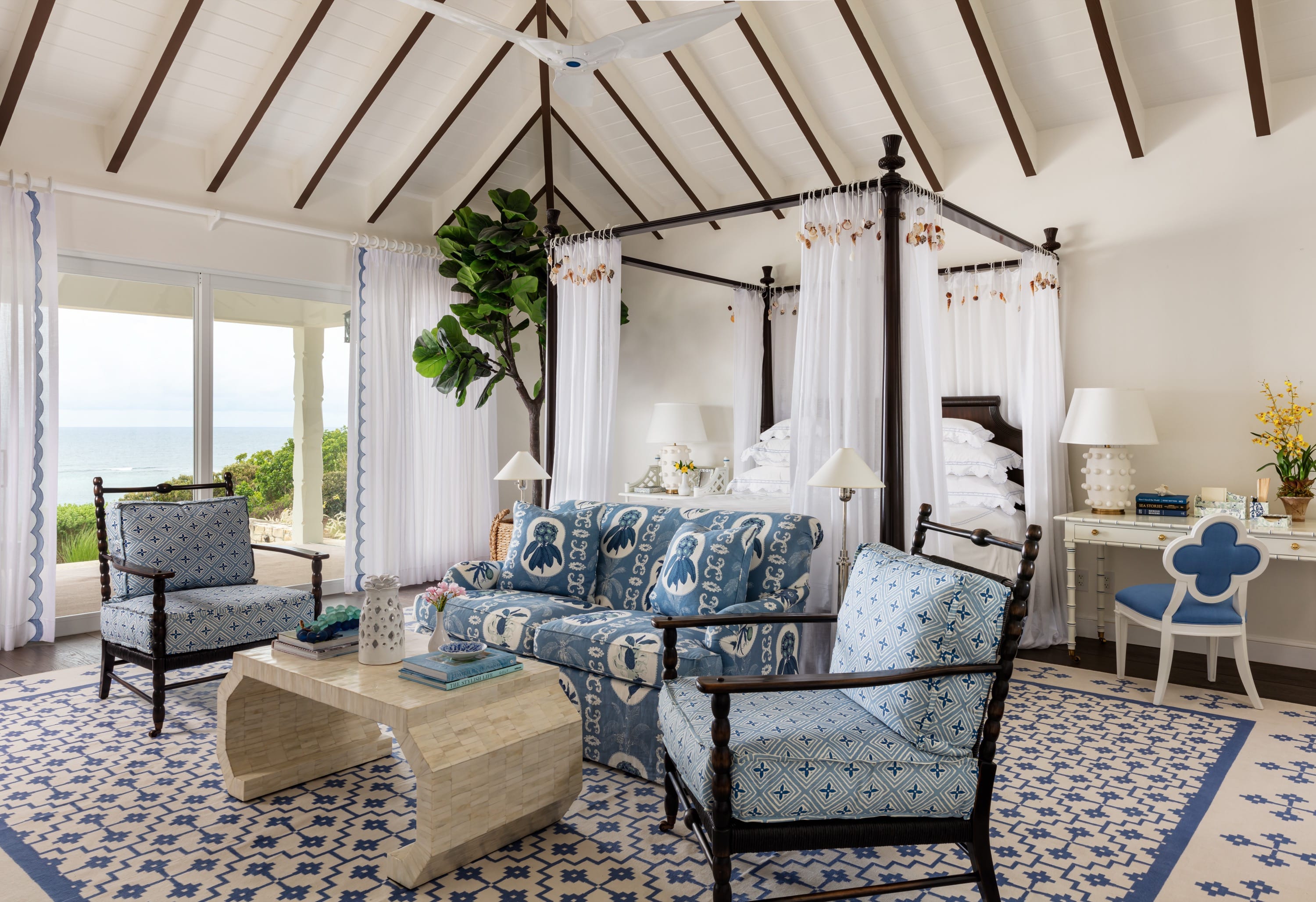 Bright bedroom with canopy bed, blue-patterned sofa and chairs, white desk, and ocean view.