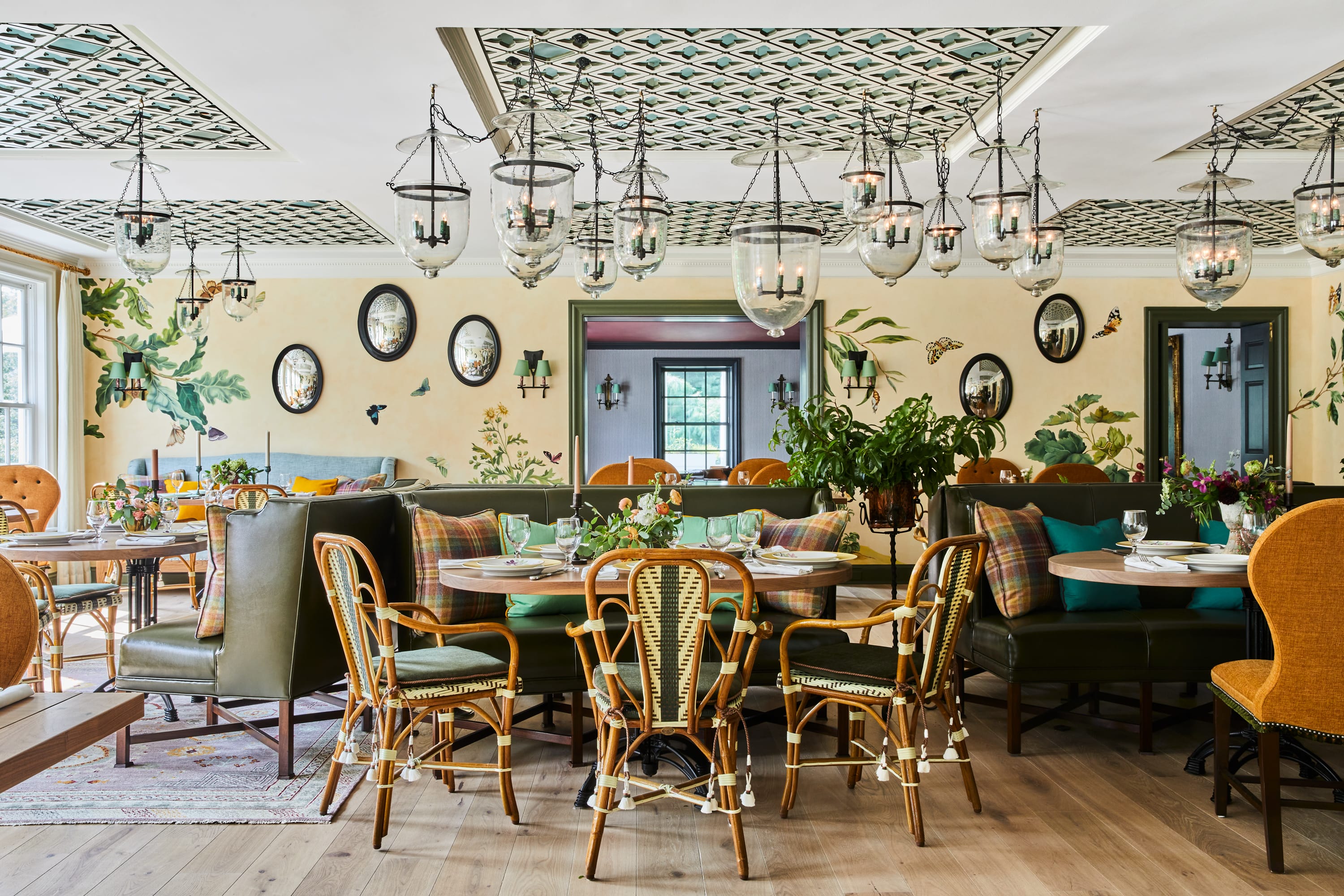 Bright restaurant interior with wooden chairs, green cushioned benches, floral wall art, and hanging glass lanterns.