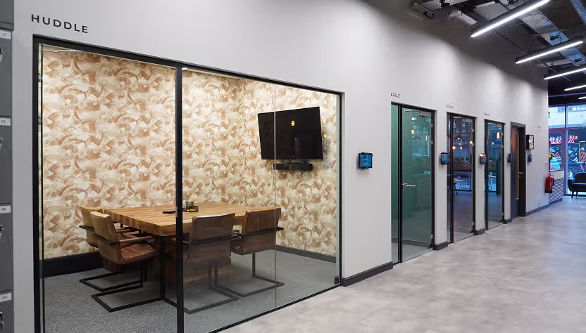 View from outside of the Huddle Meeting Room. Glass meeting room with table seating and wall-mounted display.