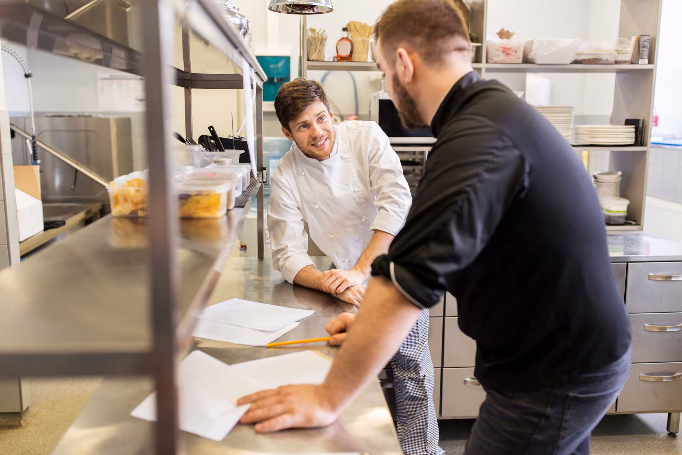 Two chefs discuss kitchen tasks while leaning over a metal counter with papers.