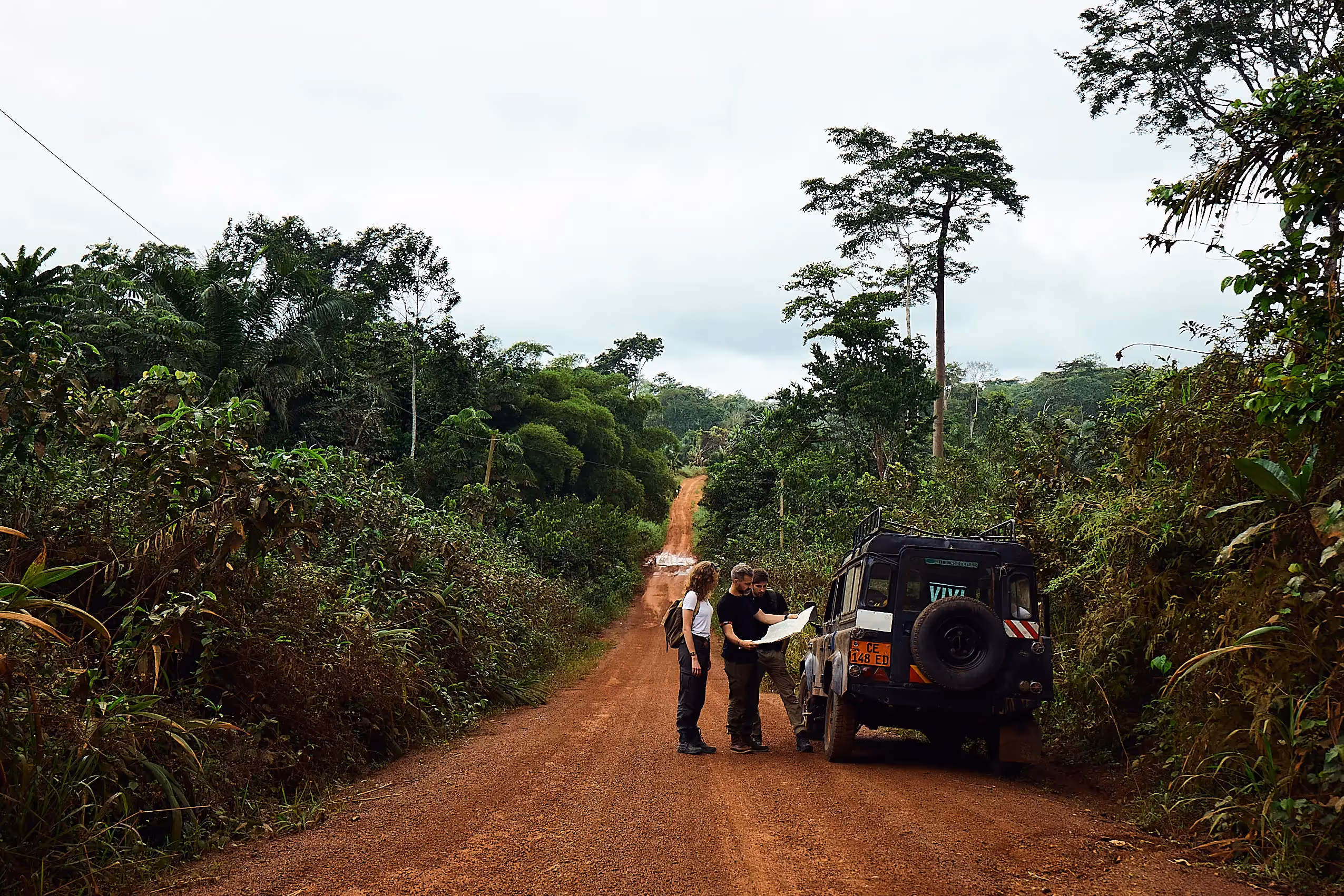 A cyclist wearing a Mondia jersey drinks from a bottle while smiling with two men, one with his arm around the cyclist's shoulders.