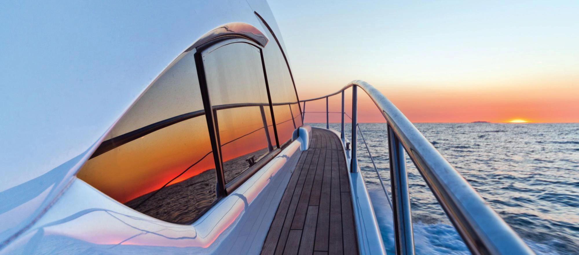 Side deck of a yacht with polished railing and reflective windows at sunset over the ocean.