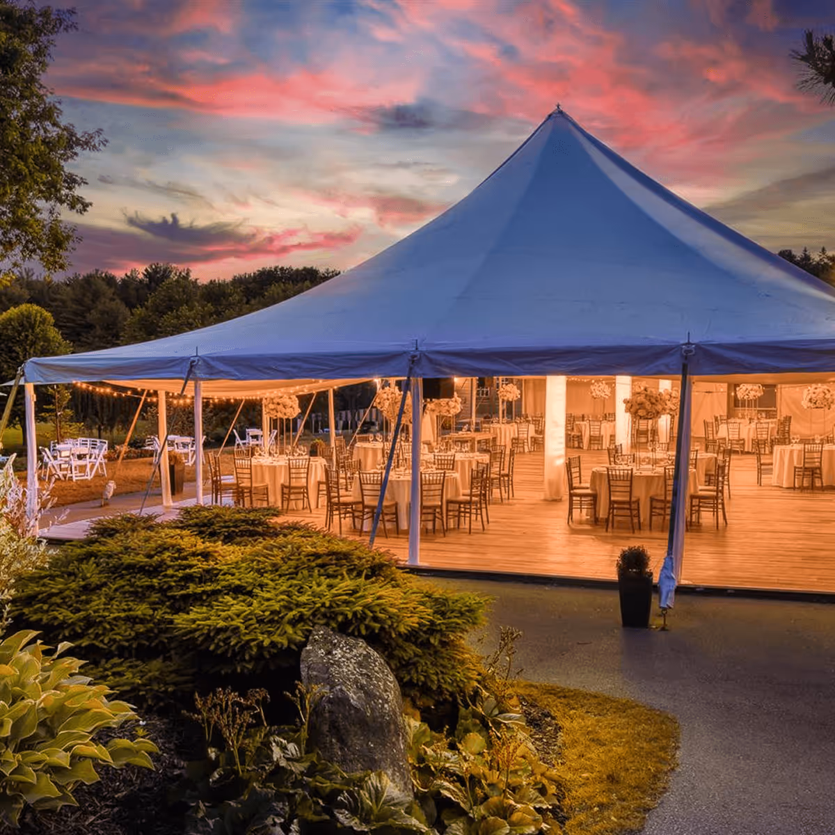 Grande tente blanche éclairée pour un événement en soirée avec tables et chaises disposées sous un ciel aux couleurs du coucher de soleil.