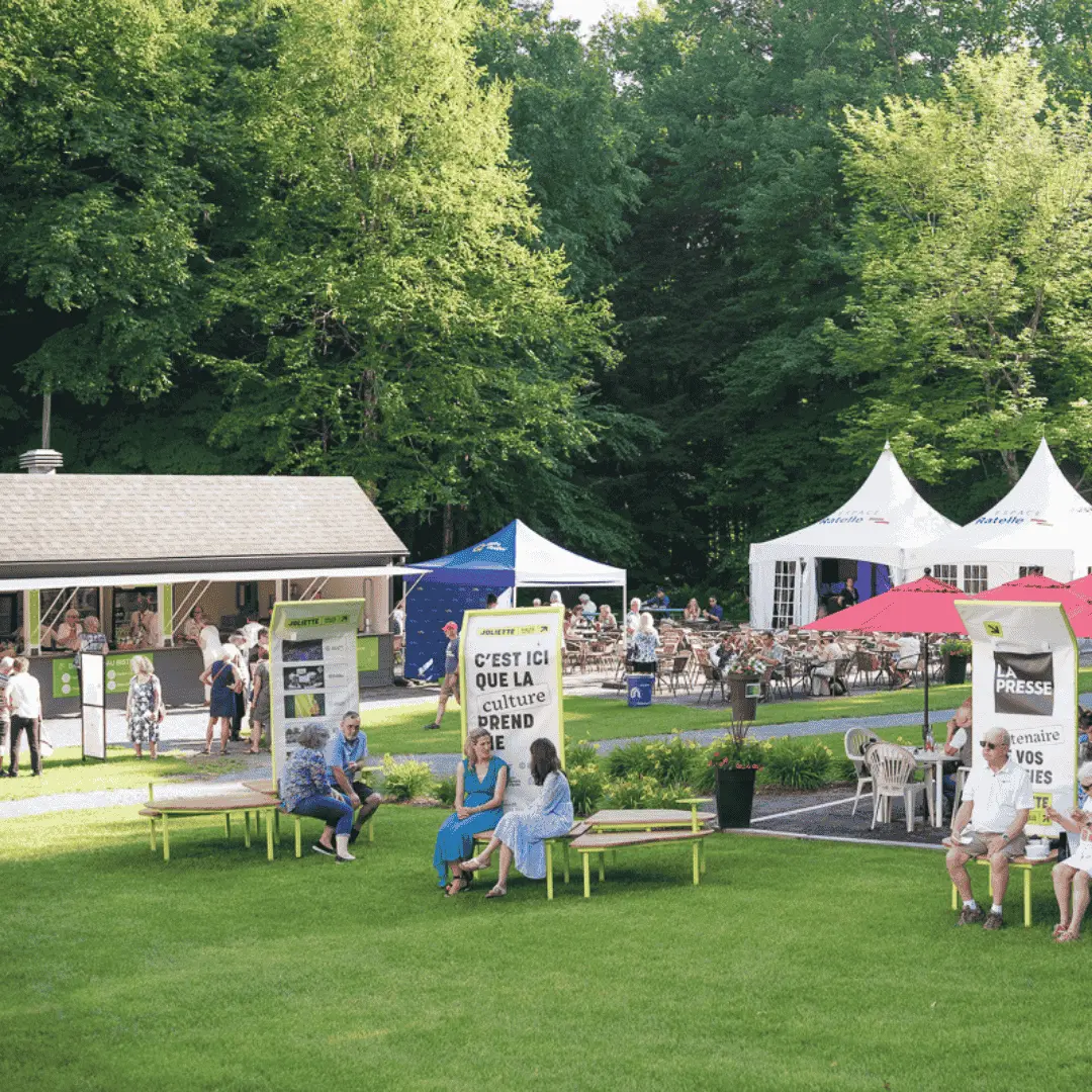 Personnes assises et discutant sur des bancs dans un parc avec des tentes, parasols et un kiosque en arrière-plan lors d'un événement culturel en plein air.