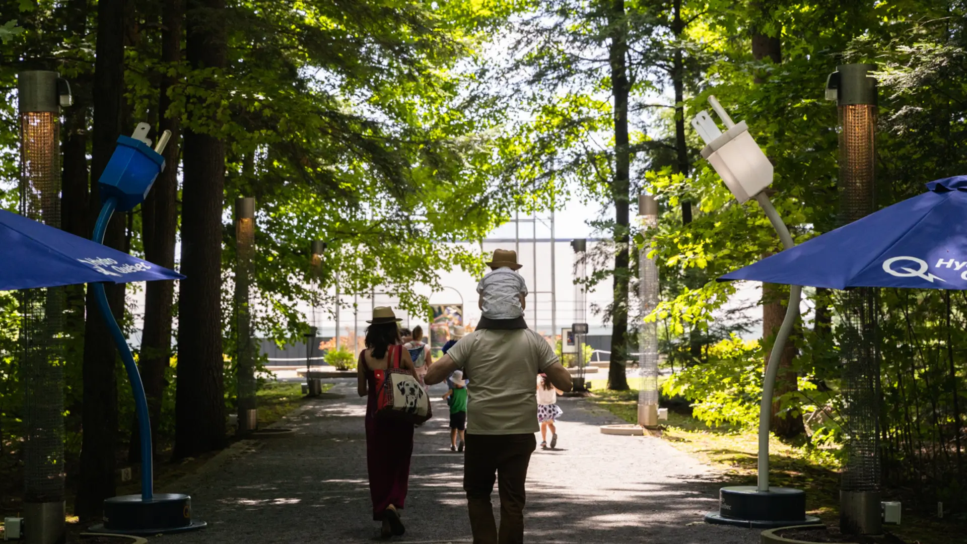 Famille marchant dans un sentier ombragé bordé d'arbres avec des sculptures de prises électriques et des parasols bleus.