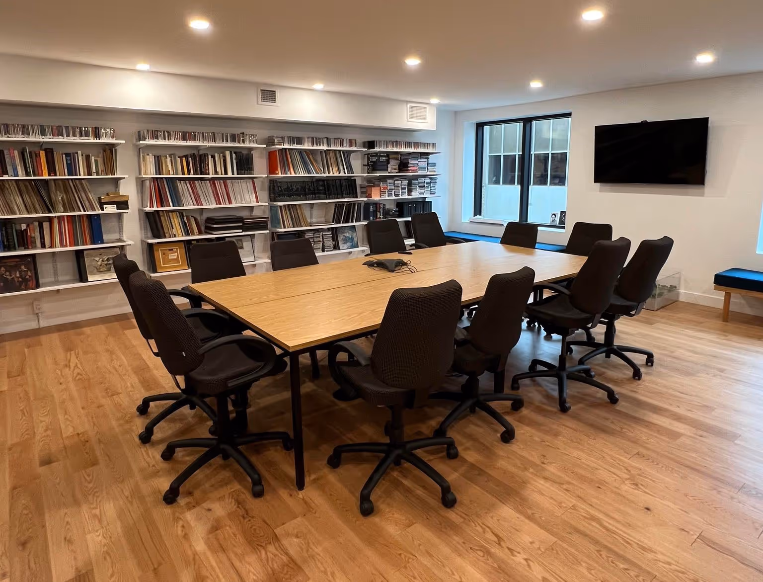 Salle de réunion moderne avec une grande table en bois entourée de plusieurs chaises à roulettes, murs avec étagères remplies de livres et vinyles, fenêtre et écran plat accroché au mur.