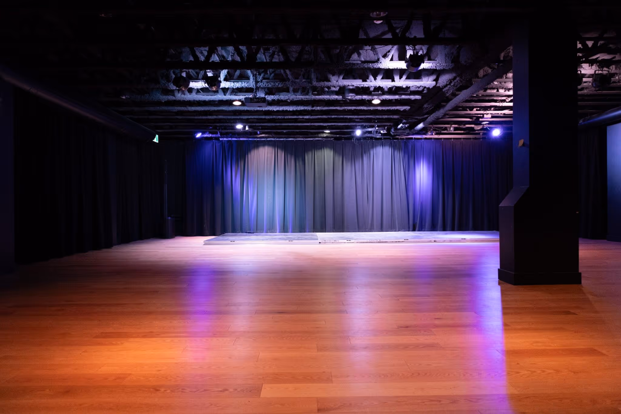 Scène vide avec rideaux noirs et plancher en bois sous un éclairage violet tamisé.