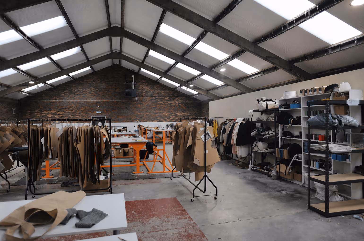 Spacious garment factory interior with hanging clothing patterns, shelves with textiles, and work tables under skylights.