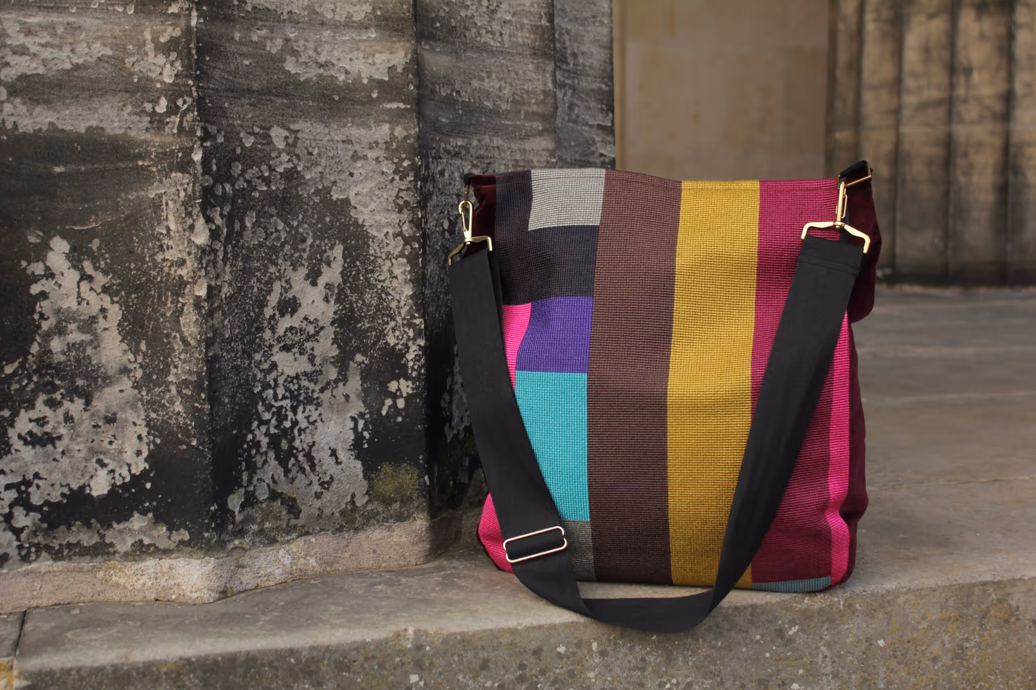 Colorful striped shoulder bag with a black adjustable strap resting against a weathered stone wall.