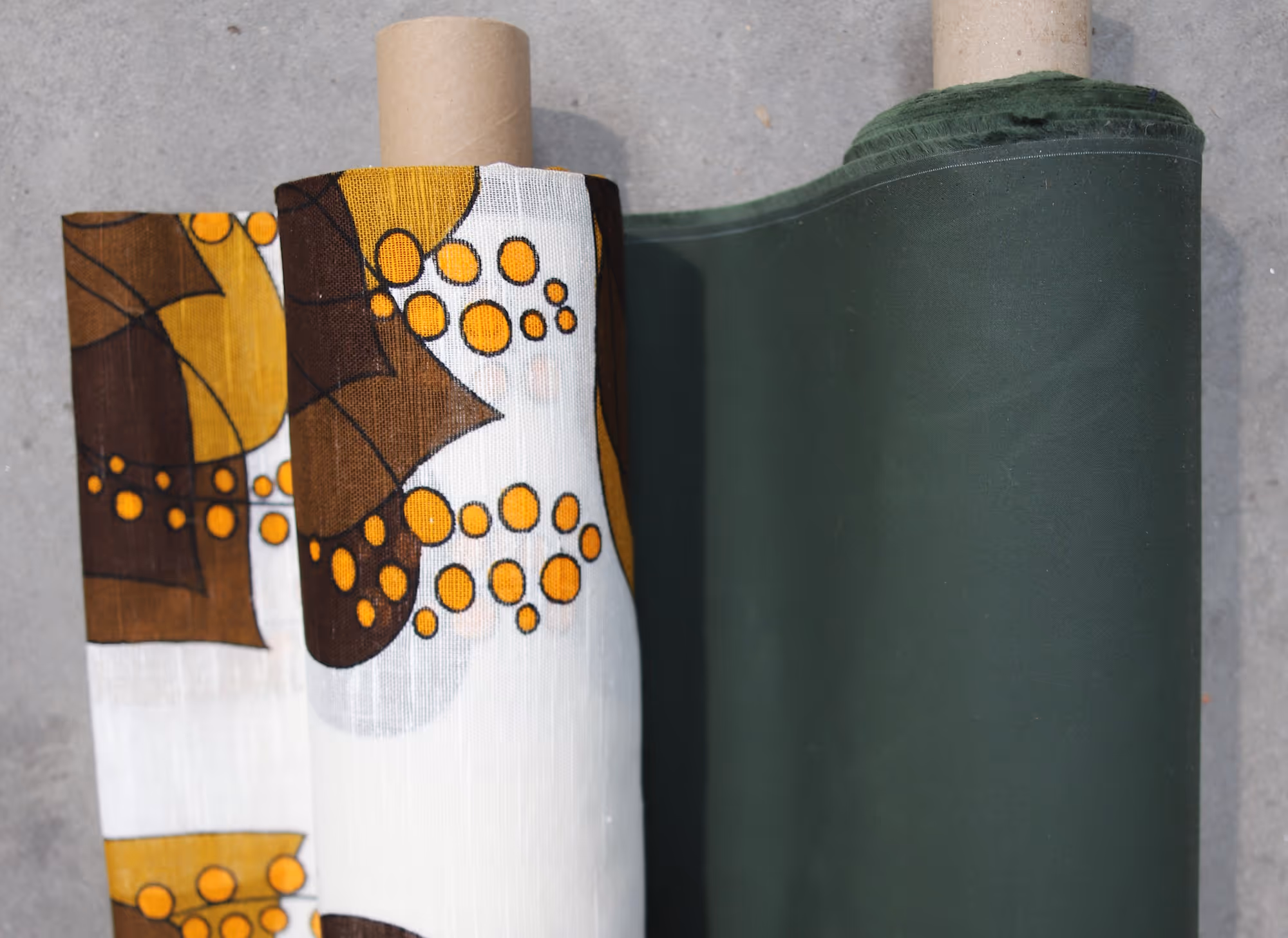 Rolls of fabric on gray surface, one with brown and yellow floral pattern on white, the other solid dark green.