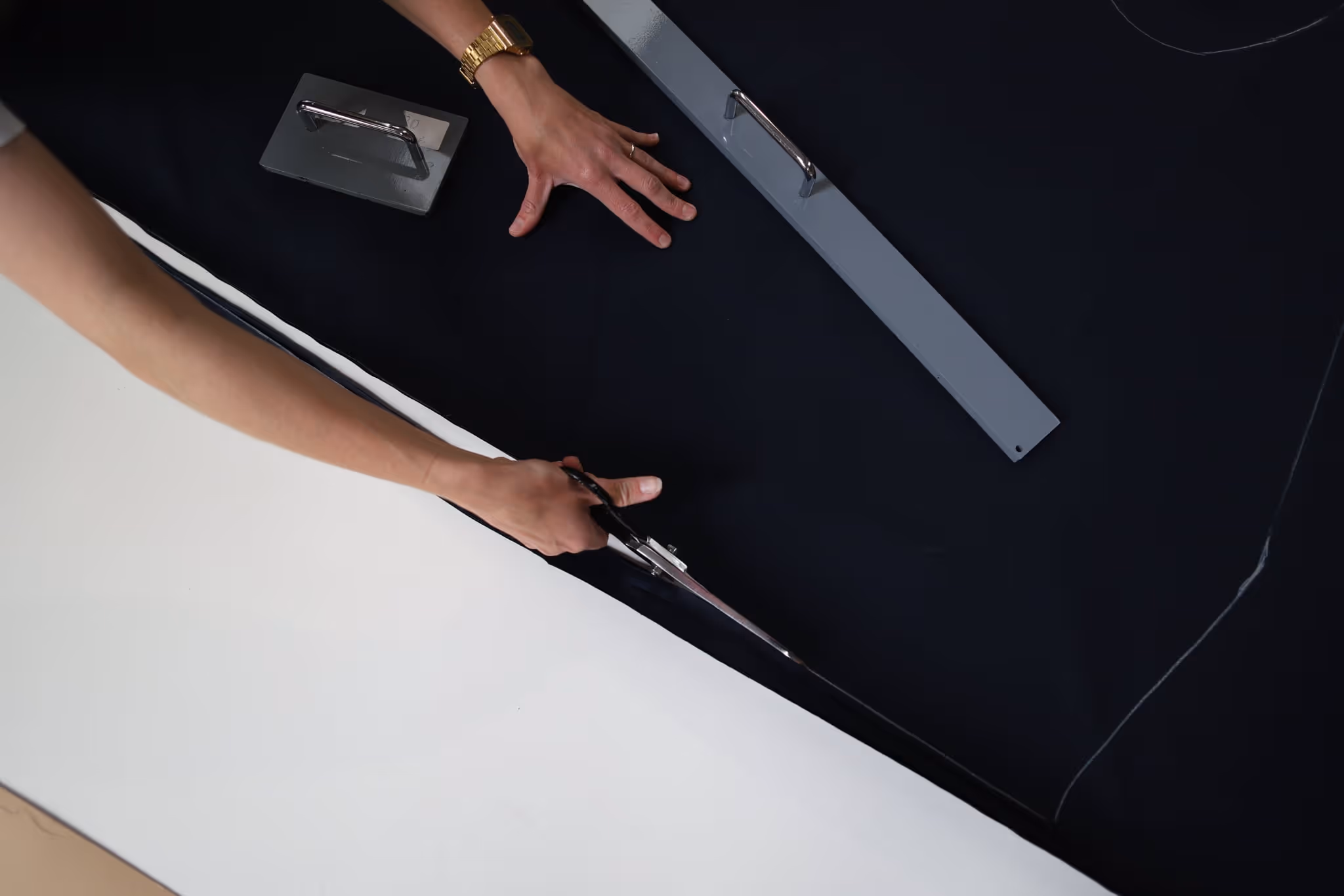 Hands cutting black fabric with large scissors using a curved ruler and pattern weights on the fabric.