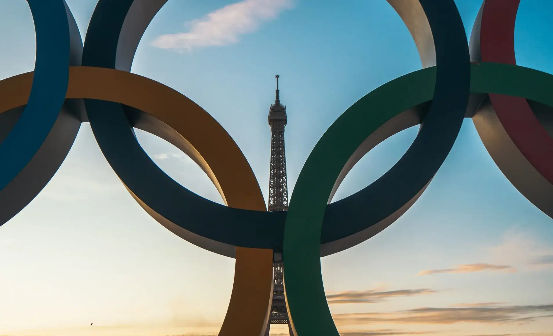 Olympic rings sculpture in Paris framing the Eiffel Tower against a sunset sky.